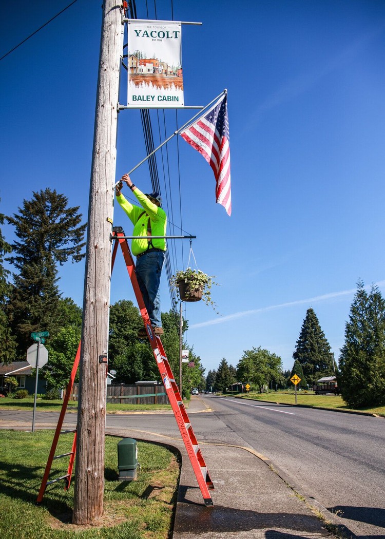 Joseph Gomulkiewicz, with Yacolt Public Works, hangs an American flag on a telephone pole above freshly watered hanging baskets along Railroad Avenue in Yacolt on Friday, May 22.